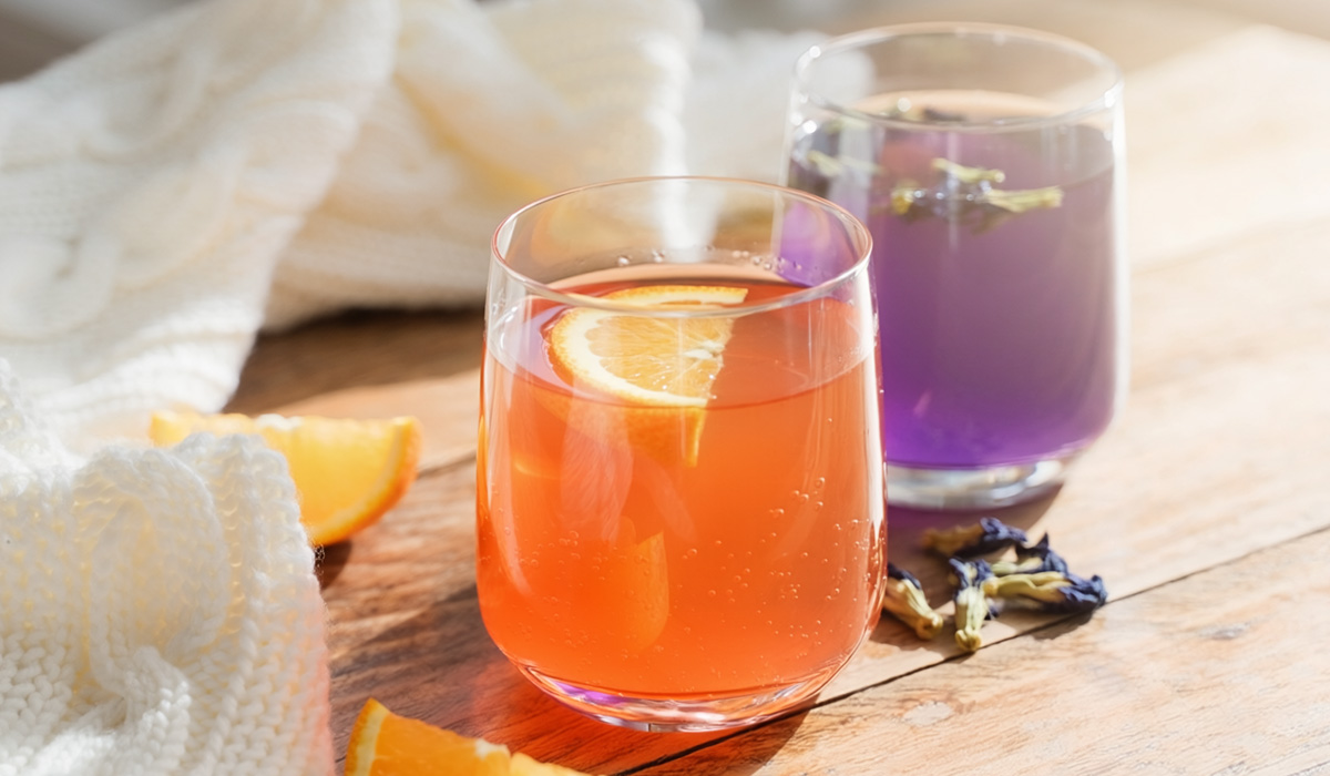 A glass of Spanish orange kombucha and purple butterfly pea kombucha on a sunlit table, offering a natural remedy for gut health winter blues. A white cable-knit blanket and orange wedges sit nearby.