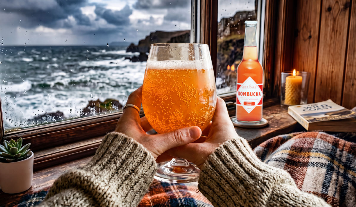 A cozy cabin window setup featuring warm winter kombucha mocktails next to a rainy ocean view.